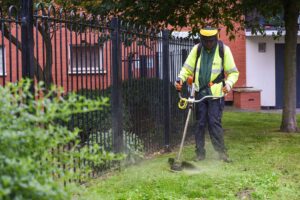 A worker in a high-visibility yellow and green jacket uses a string trimmer to cut grass along a fence. The background features a brick building and trees, while grass and plants are visible in the foreground. The worker is wearing protective gear, including a helmet and ear protection.